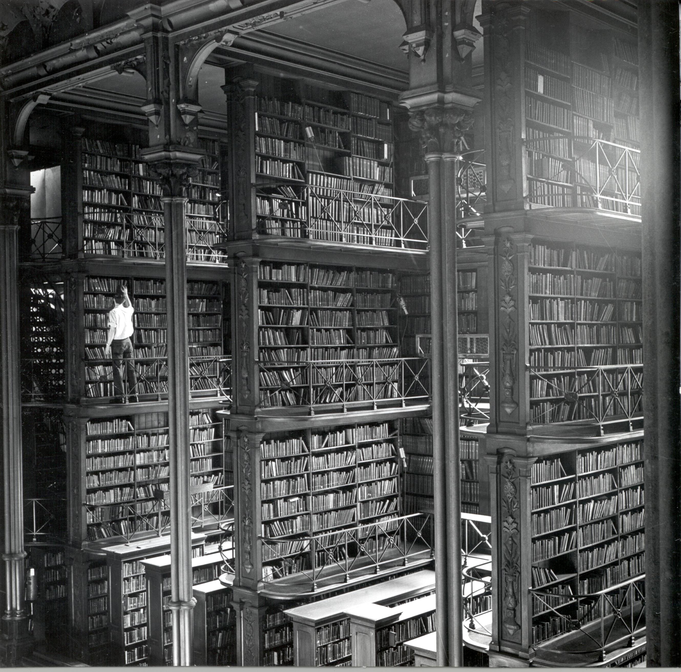 19th century image of Cincinnati public library, with four story book shelves visible in an atrium, and a man reaching for a book on the top of the third floor of one of these bookshelves.